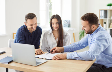 Real estate agent, mortgage broker, bank finance adviser, money loan manager gives pen to young couple clients buyers to sign paper contract after they negotiate his offer at desk with laptop computer