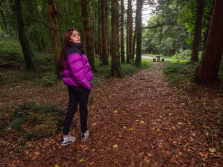 Wall Mural - Young teenager girl in a dark forest park on foot path. Travel and tourism concept. Model is with long dark hair and slim body type wearing colorful jacket and black trousers.