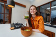 © SHOTPRIME STUDIO - Young woman in an orange shirt enjoying healthy food and green smoothie in a modern kitchen, smiles radiantly, representing healthy eating and wellness lifestyle