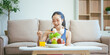 © NanSan - A happy Asian woman in sportswear sits on a sofa at a table, enjoying a salad bowl, fruits, fresh vegetables, orange juice, and energy supplements after a home fitness workout