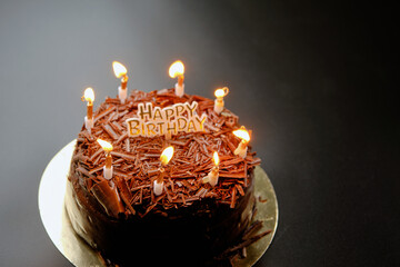 A dark black forest chocolate cake with a Happy Birthday sign sits on a black background