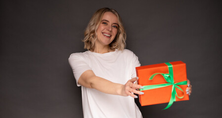 A cheerful blonde woman in a white shirt offers a bright orange gift box with a green ribbon toward the camera, smiling warmly, set against a gray background, perfect for celebration and gift themes.