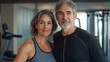© hdesert - A middle-aged couple with salt-and-pepper hair, dressed in workout gear, standing confidently in a gym setting, with fitness equipment visible in the background
