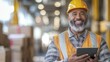 © BrightWhite - Smiling African American warehouse worker wearing hard hat and safety vest holds tablet, overseeing operations, in large warehouse environment with stock boxes in background.