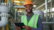 © BrightWhite - Smiling black male engineer wearing safety helmet and reflective vest, holding tablet, standing in industrial facility, monitoring processes, ensuring safety protocols, engaging with technology.