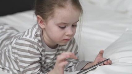 Kid studies online via smartphone. Preschooler girl does homework attentively using phone touch screen on bed in bedroom. Games applications for children education. child plays in phone app in bed.