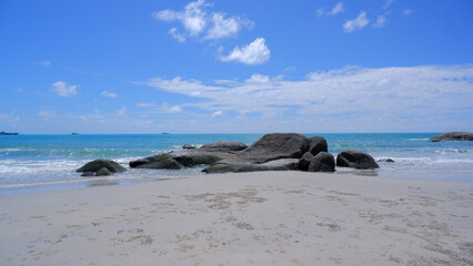 A serene beach view with large, smooth rocks scattered across the soft white sand and a few small waves crashing against the rocks.