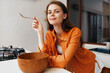 © SHOTPRIME STUDIO - Young woman smiling in a cozy kitchen, holding a spoon, wearing an orange shirt, promoting healthy eating and lifestyle