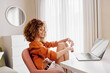 © SHOTPRIME STUDIO - A cheerful young woman with curly hair in an orange blouse and skirt, sitting at a desk while working on a laptop in a bright, modern home office