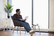 © sofiko14 - Young Indian male sitting on armchair using laptop in bright modern room. Casual attire suggests relaxed atmosphere and comfort during remote work or study.