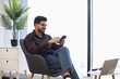 © sofiko14 - Young Indian man sitting in modern office chair using smartphone. Casual attire, relaxed atmosphere, technology interaction. Bright environment with plant and laptop adding to professional vibe.