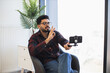 © sofiko14 - Young Indian man sitting indoors recording video content with smartphone. Wearing casual clothing, making gestures. Bright setting, natural light highlighting creative process and focused expression.