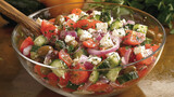 A refreshing Greek salad with tomatoes, cucumbers, red onions, Kalamata olives, feta cheese, and olive oil, served in a clear glass bowl with a wooden spoon