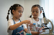 © Thanadon88 - Two young girls are looking through a microscope, one of them holding a blue spoon. Concept of curiosity and wonder as the girls explore the world of science