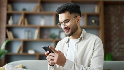 Handsome man using mobile phone sitting on sofa in living room at home. Smiling male browses social networks, does shopping in an online store, chats with a friend, writes or reads messages. Close up