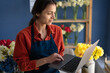 © Andrii Lysenko - woman entrepreneur working her flower shop typing on laptop standing near shelf with bouquets