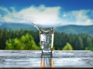 Aufkleber - Refreshing water splashes into a glass on a wooden surface, with a vibrant mountain and forest backdrop.  The scene evokes purity and nature.