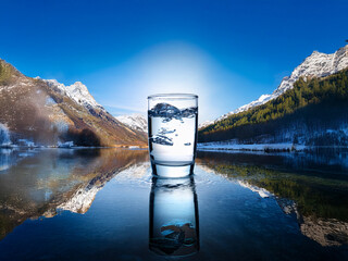 Aufkleber - A glass of pure water sits on a calm lake reflecting snow-capped mountains under a brilliant blue sky.  The scene evokes serenity and the preciousness of nature's resources.