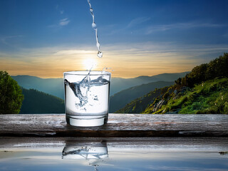 Aufkleber - Glass of water being poured, splashing, sits on wood against a scenic sunset mountain backdrop.  The scene evokes freshness and tranquility.