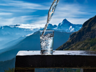 Aufkleber - Crystal-clear water pours into a glass, set against a breathtaking mountain vista under a vibrant blue sky.  The scene evokes purity and serenity.