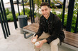 © dikushin - Cheerful handsome young man in casual attire enjoying peaceful moment on park bench in city, holding two coffee cups and laughing looking at camera, surrounded by green trees on beautiful summer day.