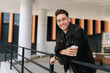 © dikushin - Portrait of happy young man holding takeaway coffee cup leaning on metal railing standing by modern building, smiling looking at camera. Serene dreaming male drinking enjoying coffee outdoors.