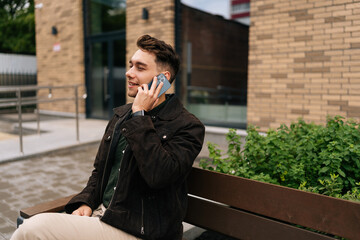 Wall Mural - Handsome bearded young man having pleasant phone conversation while sitting on urban bench near modern building in urban environment, enjoying communication. Concept of successful city lifestyle.