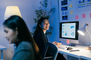 Aufkleber - Young asian businesswoman working late at night in a modern office, looking back and smiling at her coworker, with charts and graphs displayed on her computer screen