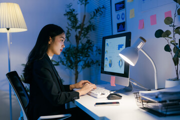 Wall Mural - Young professional woman sitting at desk typing on keyboard and using computer to analyze financial data and business charts, working late in office at night