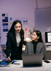 Wall Mural - Two asian businesswomen are working late in the office, analyzing data on a laptop and documents, collaborating on a project