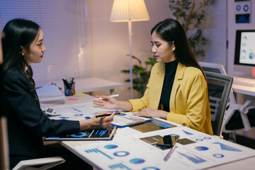 Wall Mural - Two young asian businesswomen are discussing company performance, using data charts and graphs to analyze financial results and plan business strategies in a modern office at night