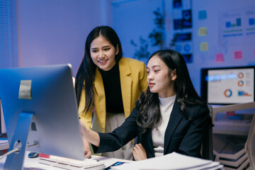 Wall Mural - Two young asian businesswomen collaborating on a project, analyzing data on a desktop computer in a contemporary office, working late at night