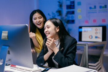 Aufkleber - Two young businesswomen are sitting at desk and looking at computer screen, celebrating successful project or good news, expressing joy and excitement in modern office at night