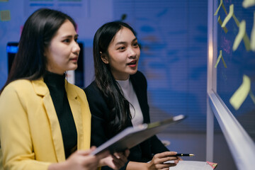 Wall Mural - Two asian businesswomen are discussing company strategy using sticky notes on a glass wall, holding clipboard and pen, collaborating on innovative solutions in a modern office environment