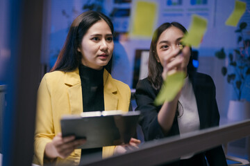 Wall Mural - Two young businesswomen are using sticky notes on a glass wall to plan their business strategy, working late at night in a modern office