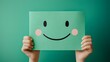 © Mitchell - Close-up shot of two hands holding a square, light green paper cutout, features a simple hand-drawn smiley face with black eyes and a curved smile, accented by pink cheek circles.