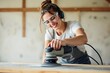 © ThomasLENNE - Cheerful female artisan in protective glasses using special electric orbital sander to smoothen wooden planks while working in countryside on summer day