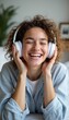 © natakot - A cheerful young woman with curly hair smiles brightly while wearing white headphones, immersed in her favorite tunes. Her relaxed demeanor and cozy attire create a warm, inviting atmosphere, perfect
