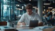 © Mrammon - A young professional writing in a notebook while working on a laptop in a modern coworking space.