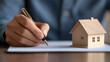 © praewpailyn - Close up of hands signing property insurance document with model house, symbolizing home ownership and security