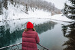 © Aleksey - A cheerful woman, wearing a vibrant red hat, stands beside a serene and peaceful lake that is beautifully surrounded by glistening white snow, fully enjoying the wonderful winter scenery