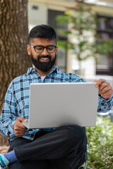 Indian bearded man freelancer working online distant job with laptop browsing website chatting sitting outdoors. Hispanic guy on city street looking at notebook screen send messages watching movies
