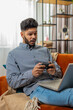© Andrii Iemelianenko - Excited Indian Arabian man playing video game on laptop spending leisure time at home sofa couch. Happy Hispanic freelancer guy playing video games with joystick controller on computer pc. Vertical