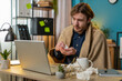 © Andrii Iemelianenko - Sick ill young businessman suffering from cold flu or allergy working on laptop at home office. Freelancer bearded man wrapped in blanket blows sneezes wipes snot into napkin. Coronavirus quarantine.