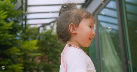 A young Asian child girl enjoying a sunny day outside surrounded by floating soap bubbles in Garden lush greenery in background, capturing the innocence and joy of childhood in natural light.