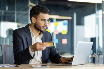 Sticker - Latin American businessman sitting at desk in office, conducting online transactions on laptop with a credit card in hand, highlighting modern business and technology.