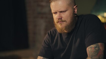 Serious tattooed man surfing internet by laptop at home in evening, portrait. Brawny male person typing messages in social networks, online shopping or remote work, internet addiction of modern people