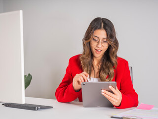 Young caucasian office worker businesswoman using tablet sitting desk at office shocked happy face expression. Wear formal red jacket, 20s female employee. Using application concept image.