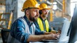 © Vilaysack - Two construction workers in hard hats focus on their computers at a job site, collaborating on project management and planning.