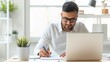 © Vilaysack - A focused man works at a laptop while taking notes, surrounded by a plant and bright natural light in a modern workspace.